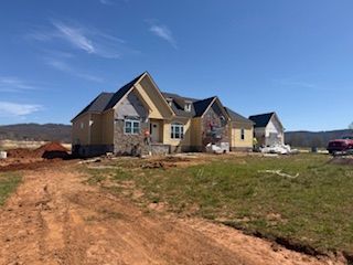 A large, single-story house under construction with yellow siding, stone accents, and a dark shingled roof on a sunny day. 