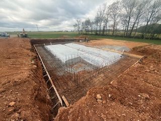 A rectangular concrete foundation with vertical steel rebar, set in a deep dirt excavation under a cloudy sky. 