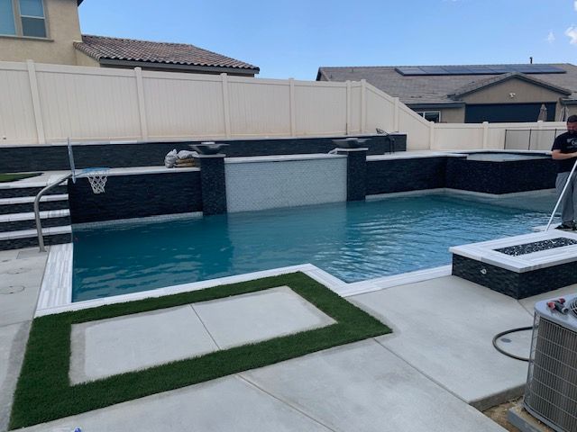 Swimming pool with waterfall, spa, and fire pit in a backyard with a white fence.