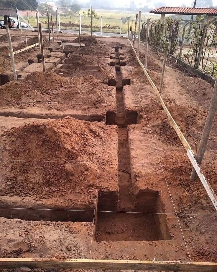 Construction site with trenches and post holes dug in reddish-brown earth for a building foundation.