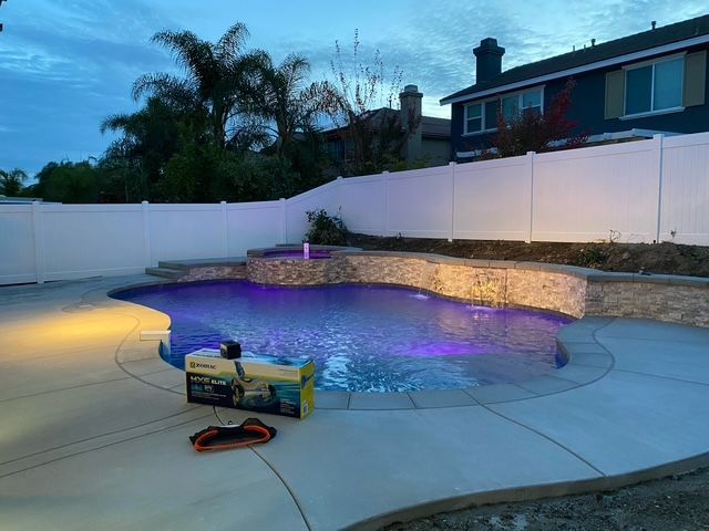 Backyard pool at dusk with a waterfall, purple underwater lights, and a white fence.