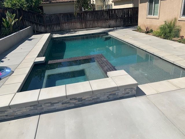 Rectangular pool with attached spa, surrounded by concrete. Backyard setting with a fence.