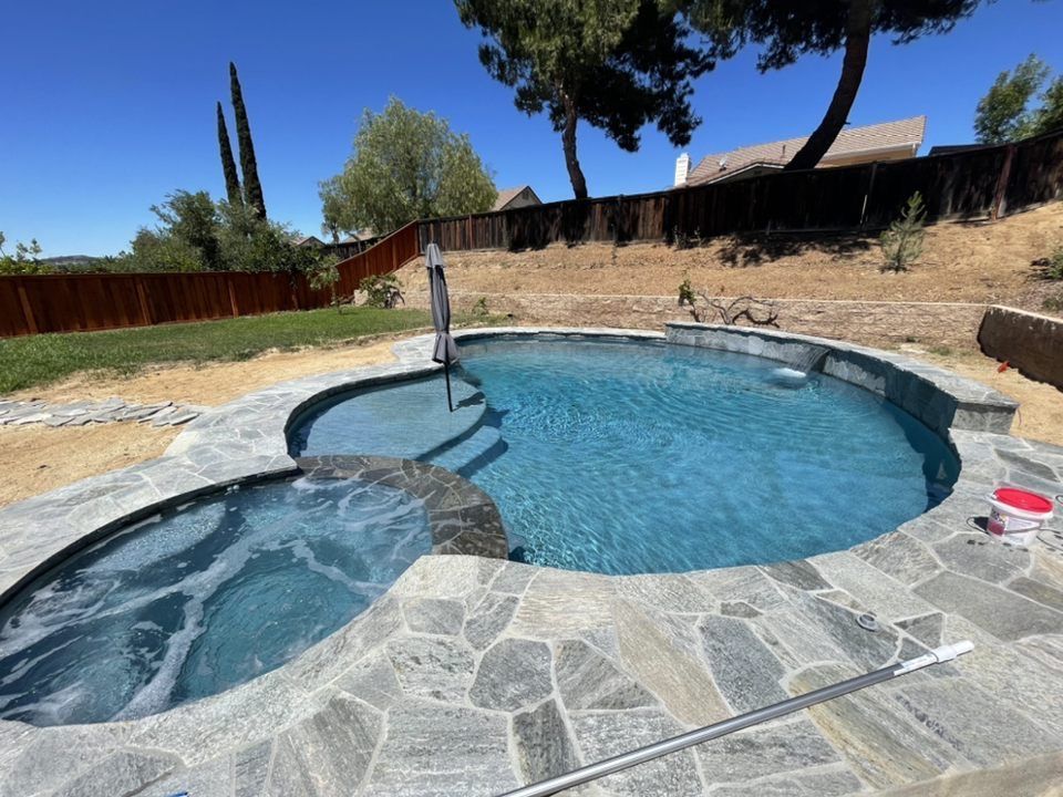 Pool and spa with gray stone surround, set in a sunny backyard with a wooden fence.