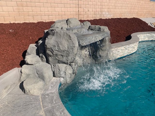 Water cascades from a rock formation into a turquoise pool, and red mulch and a brick wall are in the background.