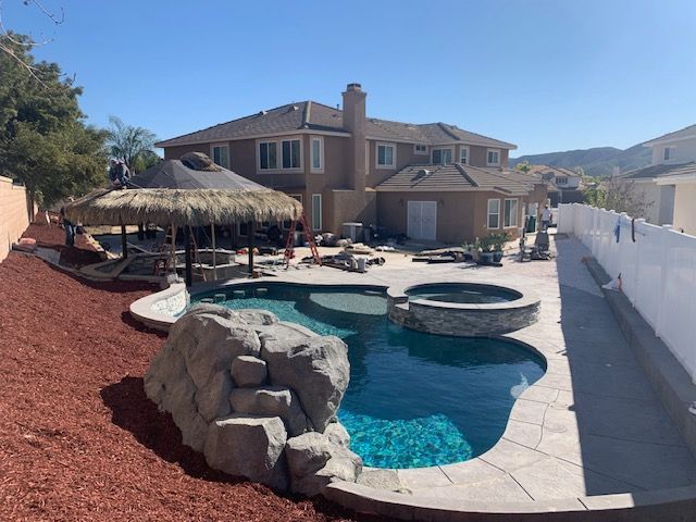 Backyard with a pool, hot tub, and tiki hut. House in background, red mulch, and blue water.