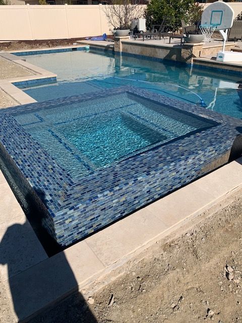 Square blue-tiled hot tub with steps in a larger pool, tan concrete patio.
