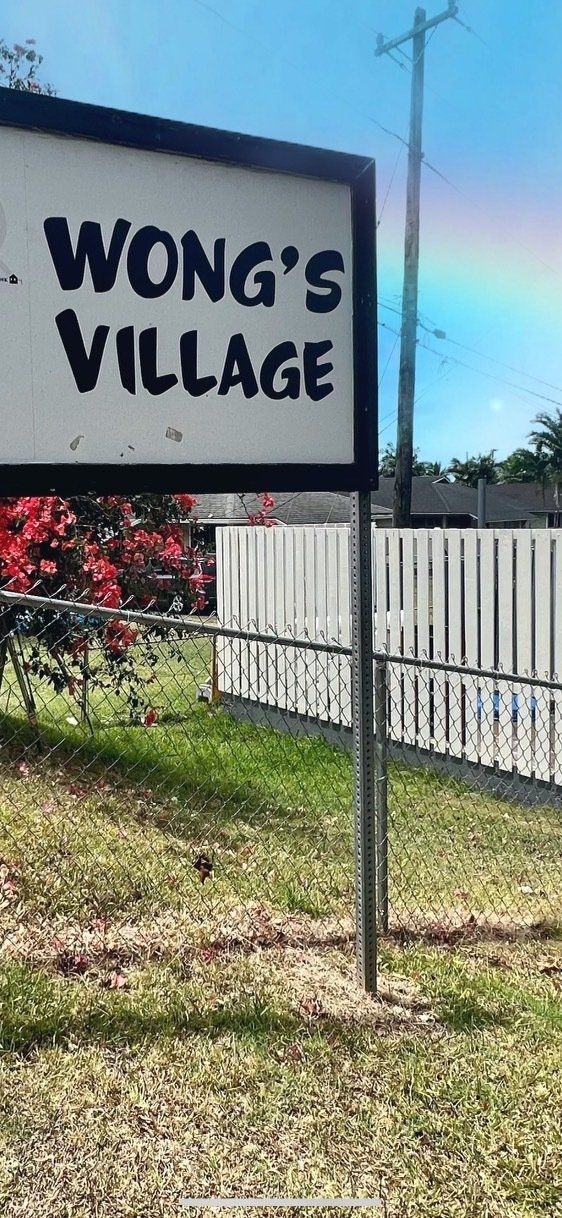 Sign for Wong's Village, set outdoors with a rainbow and white fence.