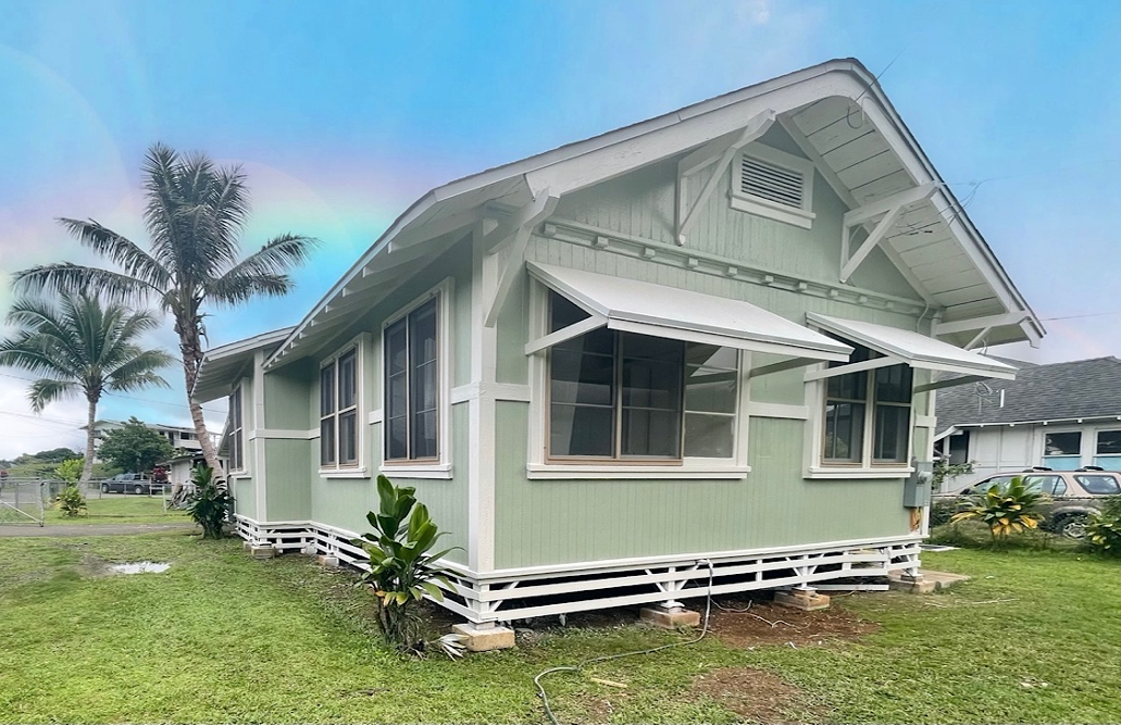Green and white cottage with white awnings, palm tree, and a rainbow in the blue sky.