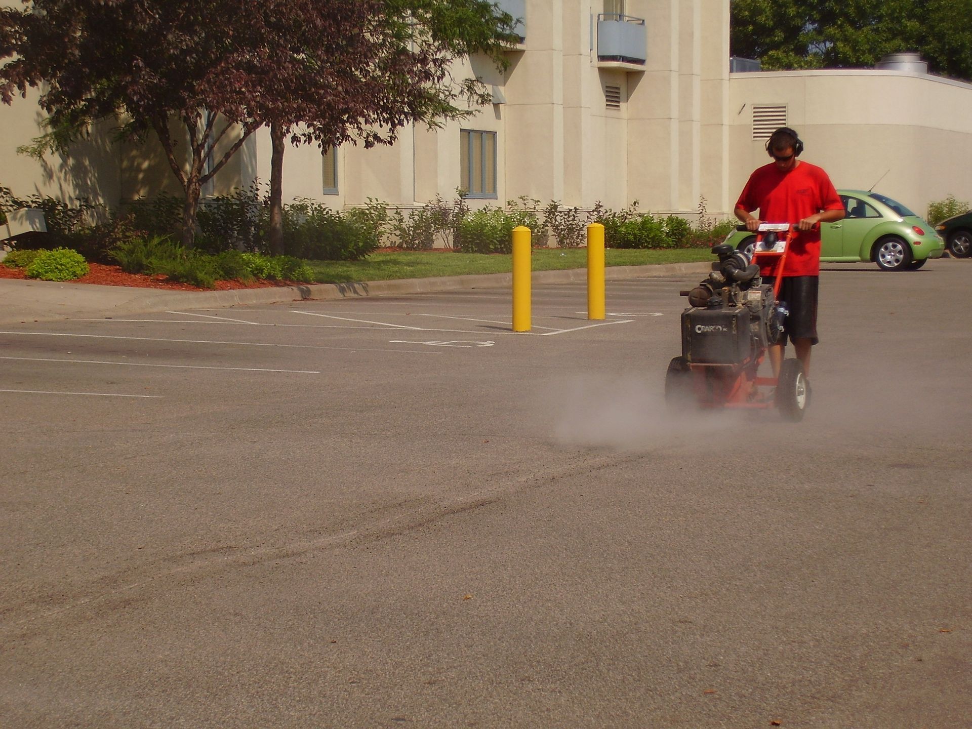 Person in red riding a lawn mower through a parking lot, kicking up dust near a building.