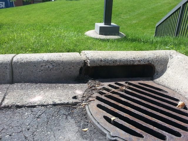 Storm drain grate by curb with a concrete sidewalk and green grass lawn nearby