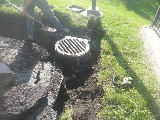 Damaged sidewalk with an open manhole and exposed dirt beside a grassy yard