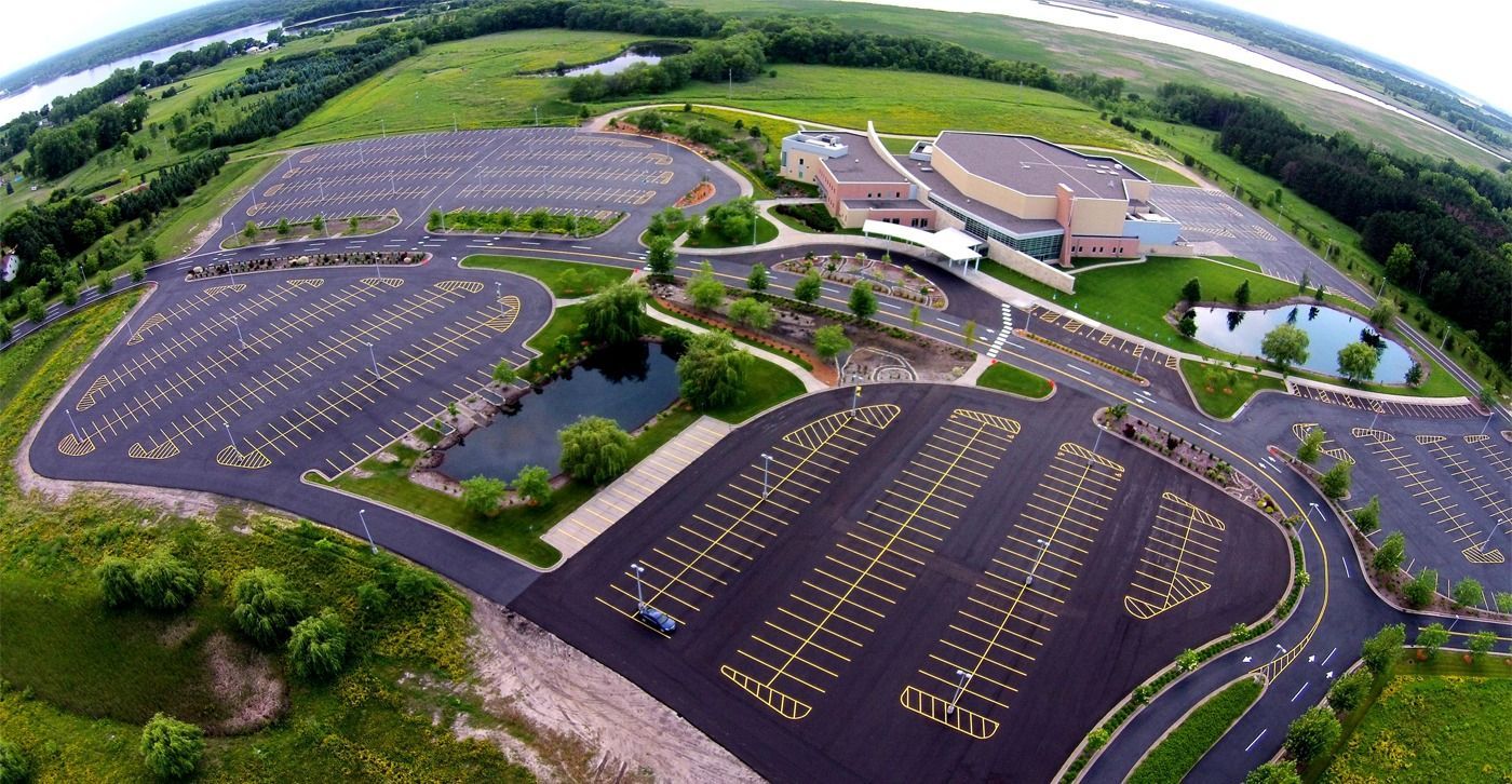 Aerial view of a large mostly empty parking lot beside a building, surrounded by trees and greenery.