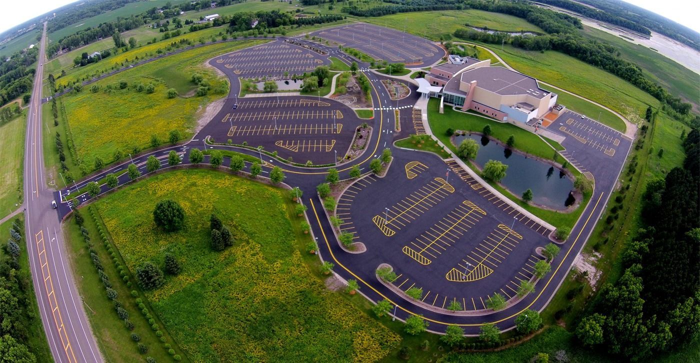 Aerial view of a modern building with purple parking lots, surrounded by green fields and trees.