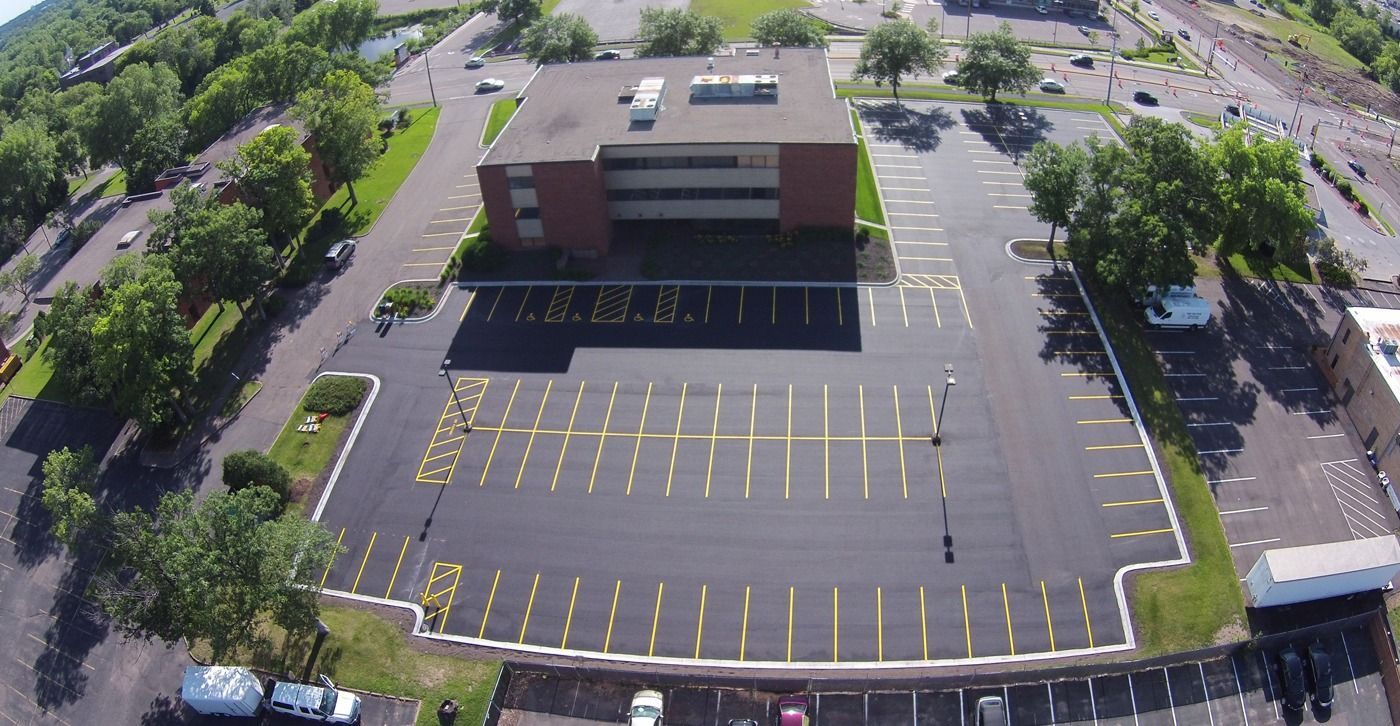 Aerial view of an empty parking lot beside a brick building with yellow-lined spaces and surrounding trees
