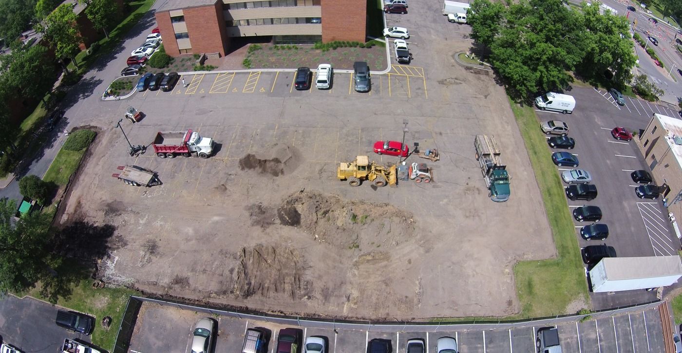 Aerial view of a large dirt lot with construction equipment, parked cars, and apartment buildings around it