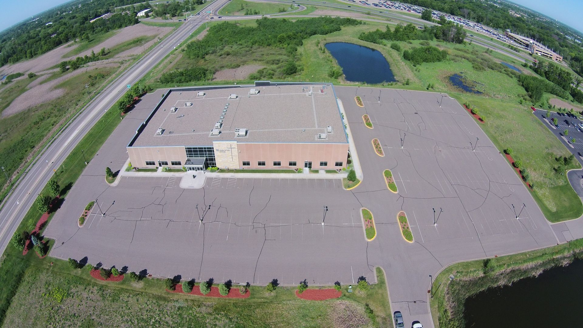 Aerial view of a large tan commercial building with a mostly empty parking lot and surrounding greenery.