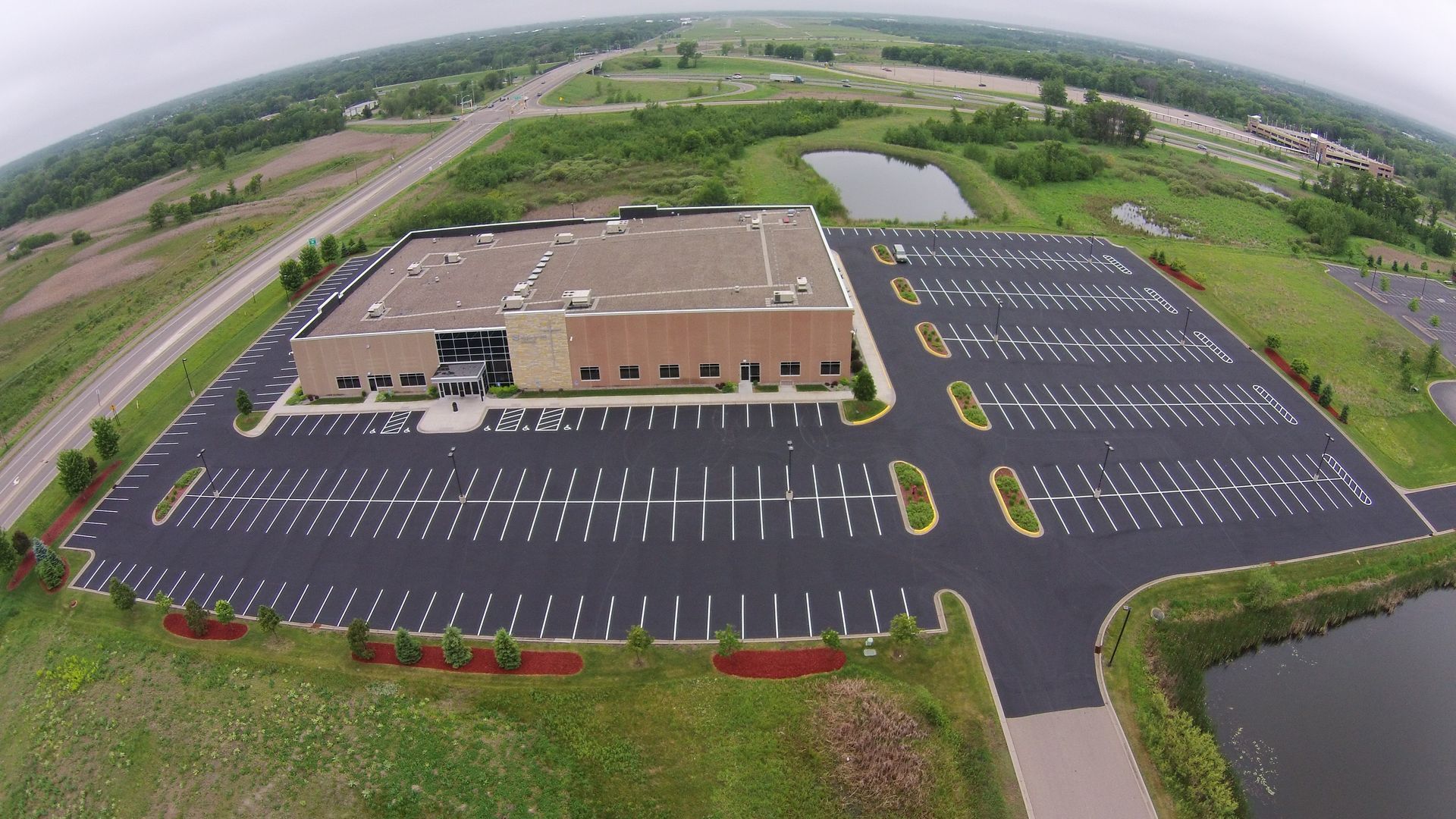 Aerial view of a large brick building with an empty parking lot and surrounding greenery.