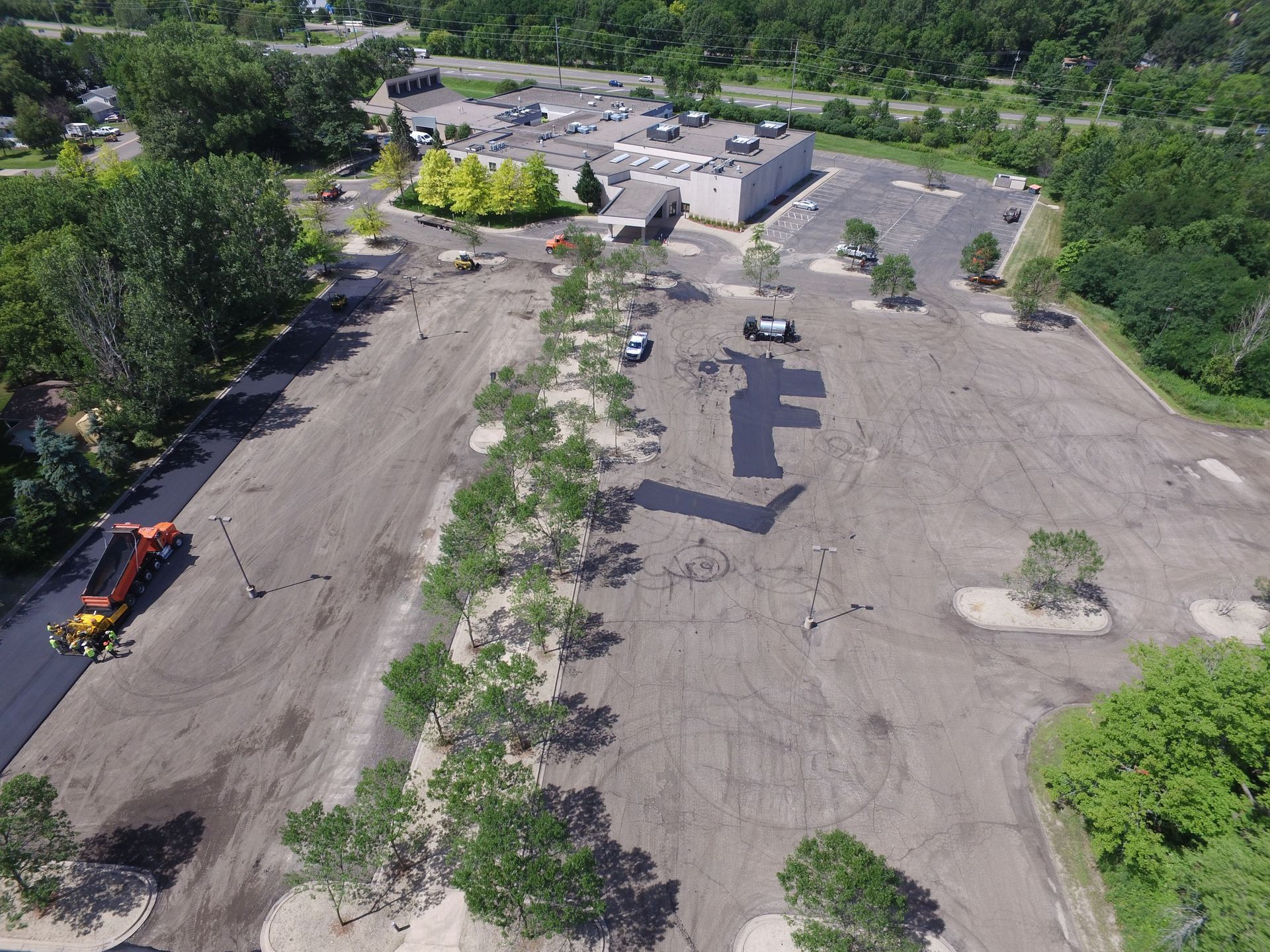 Aerial view of a large empty parking lot beside a building, with a few cars and surrounding trees.