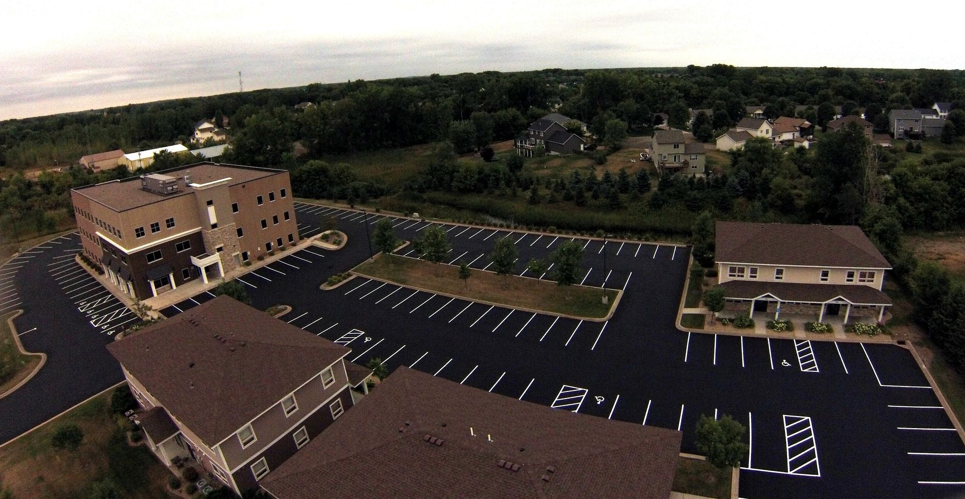 Aerial view of a commercial building complex with a large empty parking lot and surrounding trees