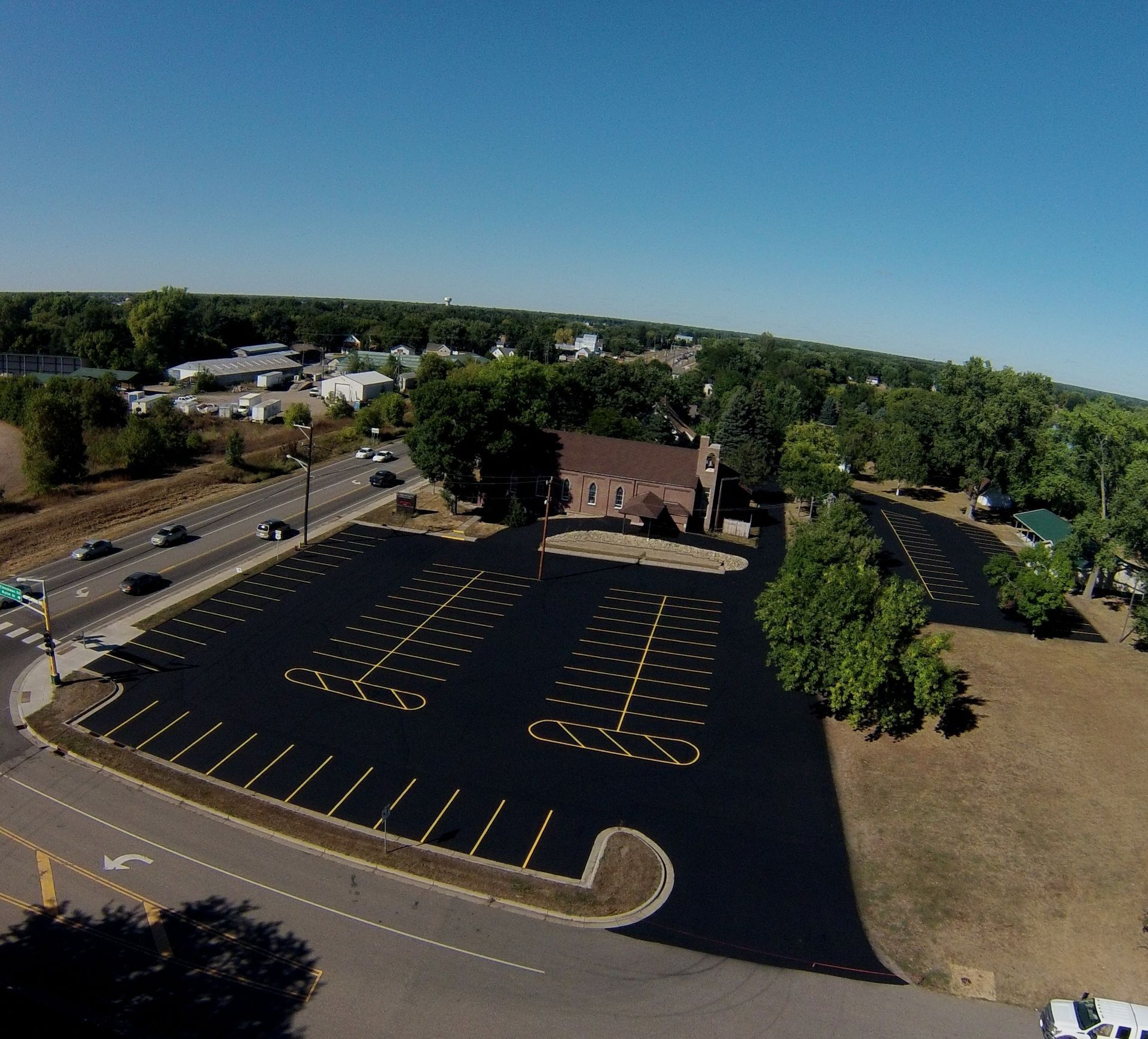 Aerial view of a mostly empty blacktop parking lot beside trees and a building on a sunny day