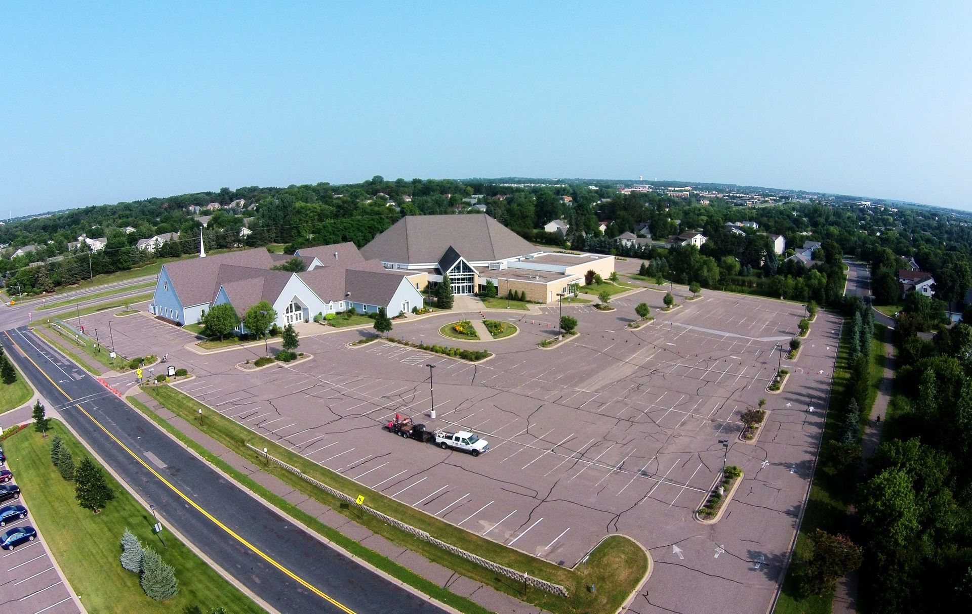 Aerial view of a large church and mostly empty parking lot beside a road and trees.