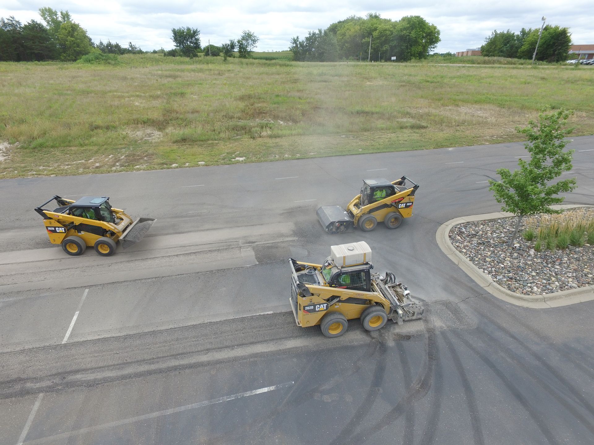 Three yellow skid-steer loaders on a paved lot, one kicking up dust near a curb and tree.