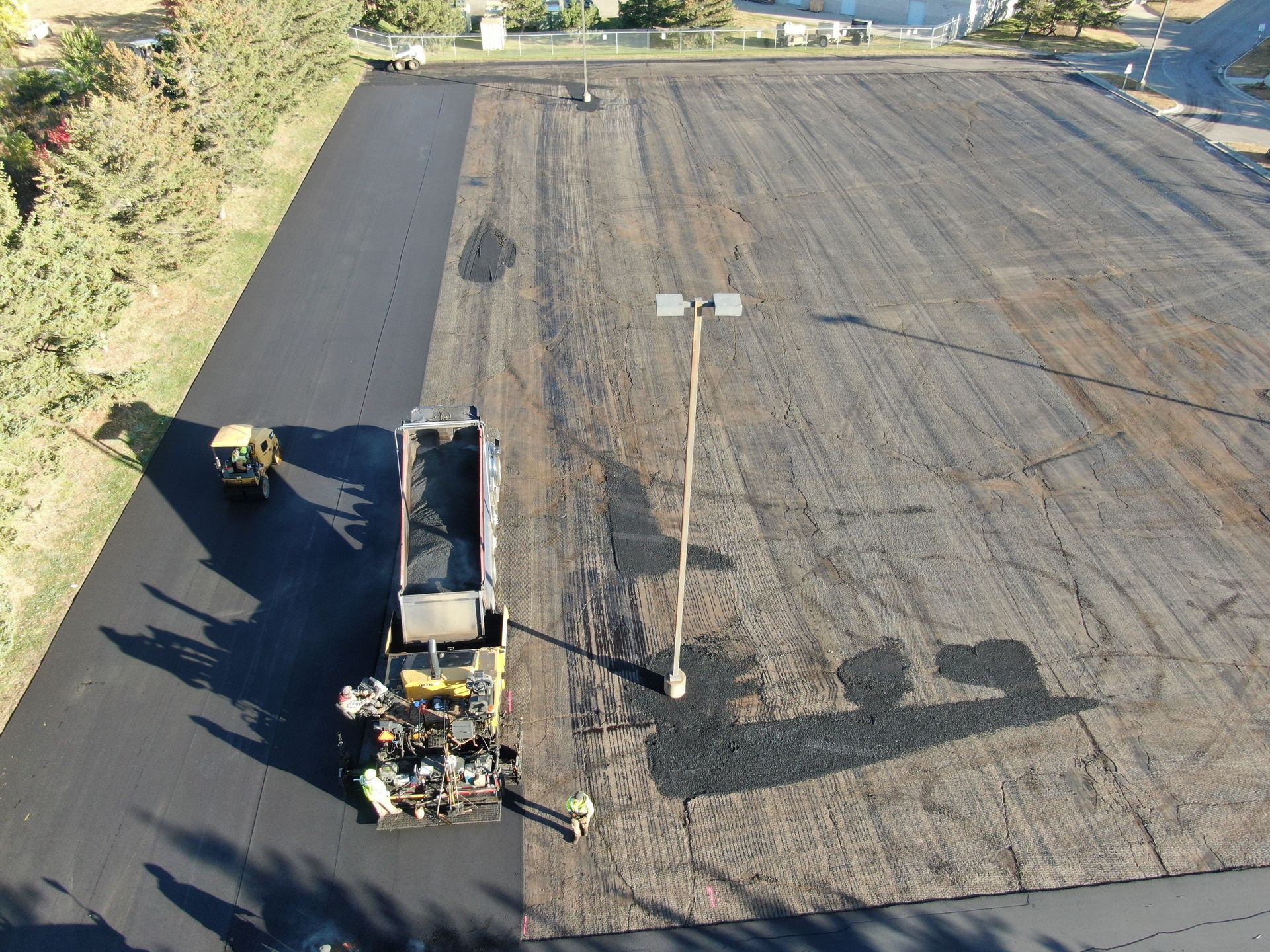 Road paving crew working on a large asphalt surface beside a dirt edge, viewed from above.