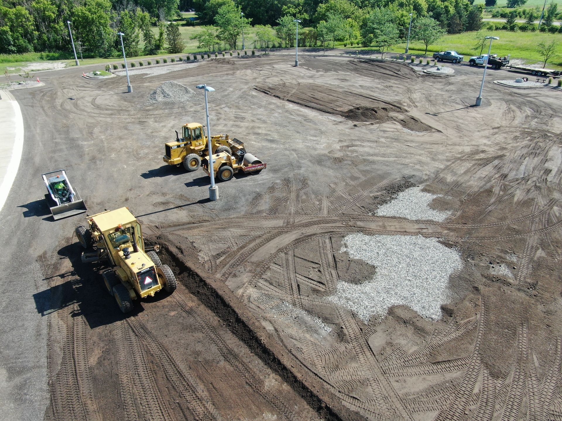Construction site with yellow heavy machinery grading a large dirt lot near trees and pavement.