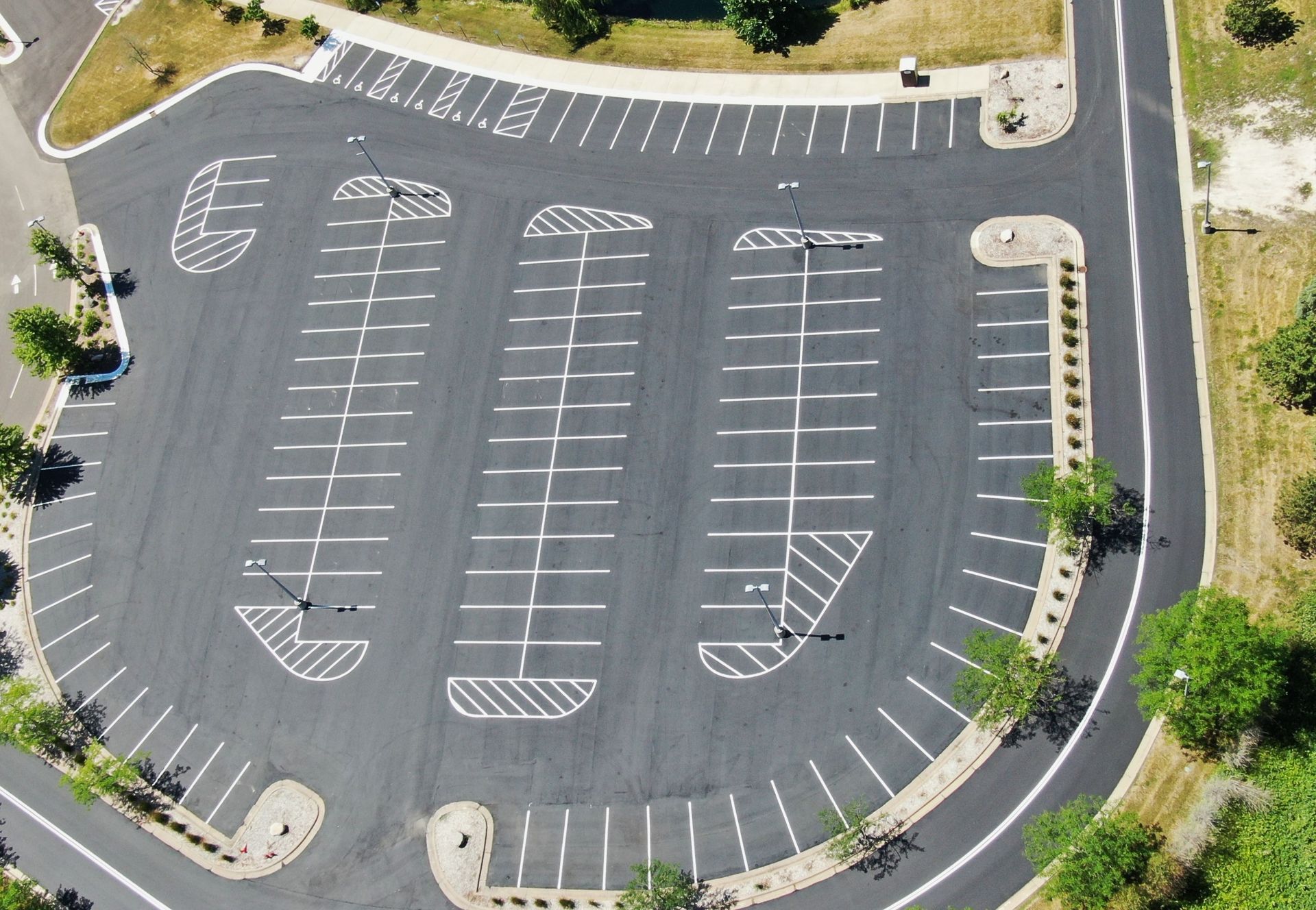 Aerial view of an empty parking lot with marked spaces, curving driveways, and surrounding grass and trees