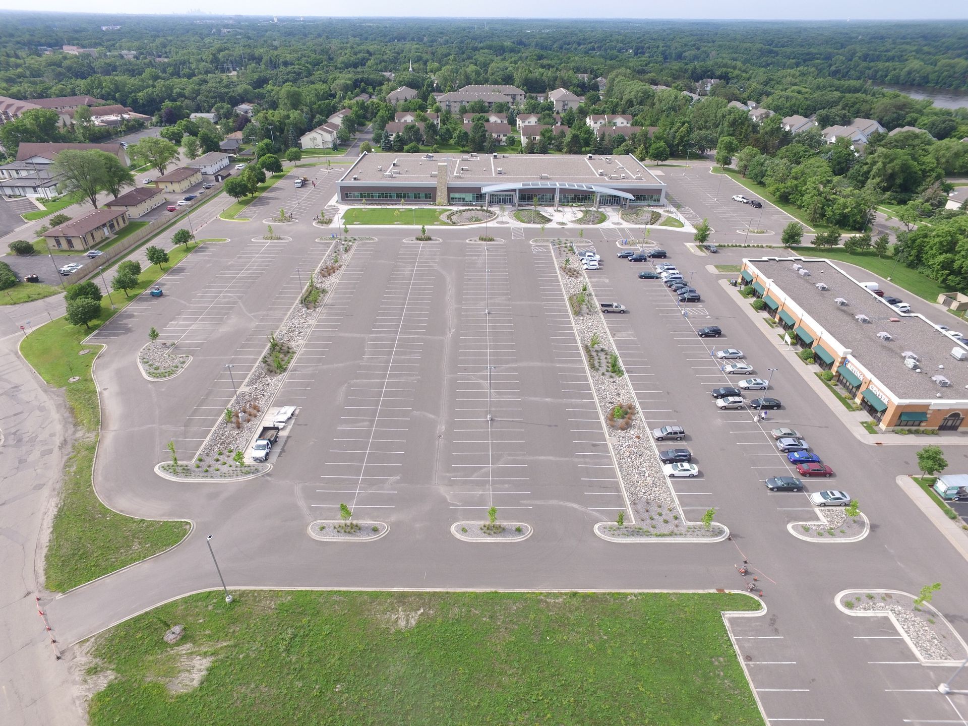 Aerial view of a large mostly empty shopping center parking lot with a few parked cars and surrounding trees.