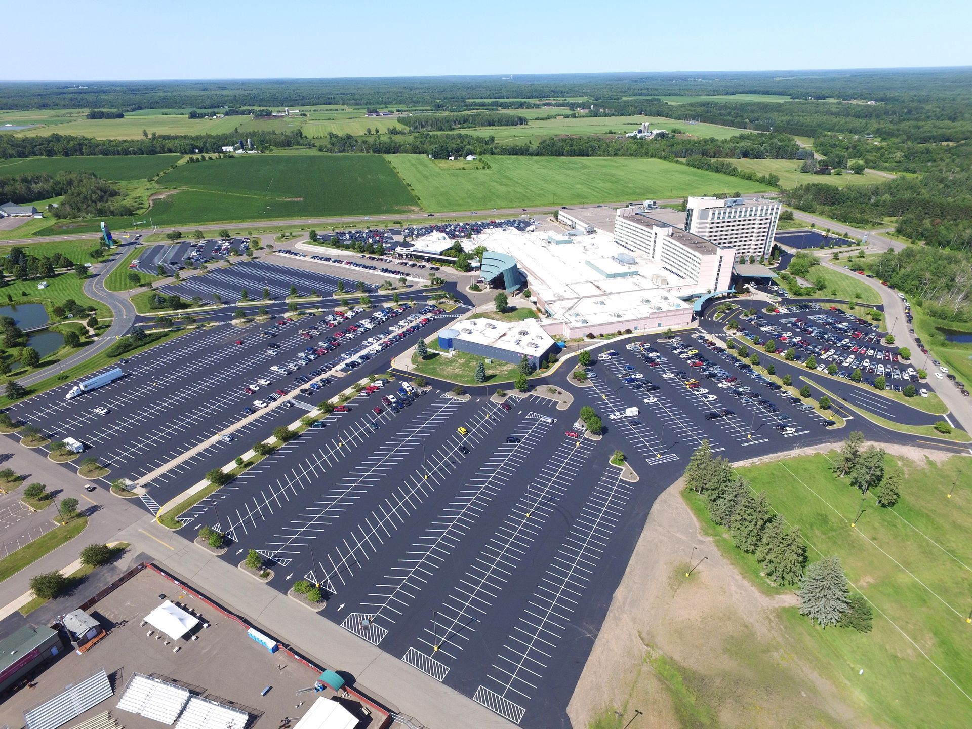 Aerial view of a large parking lot beside a white commercial complex in a green rural landscape.