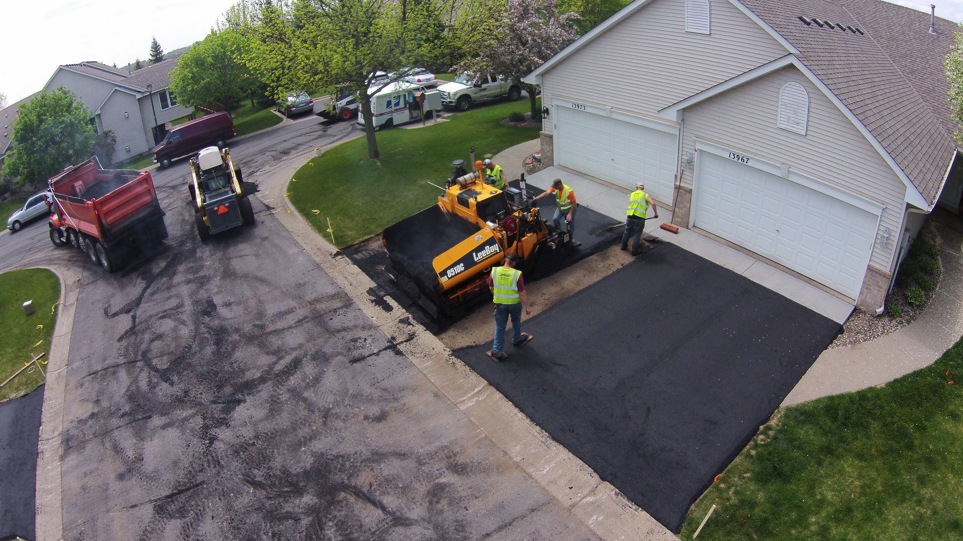 Road crew paving a black driveway beside a white garage, with yellow machinery and workers