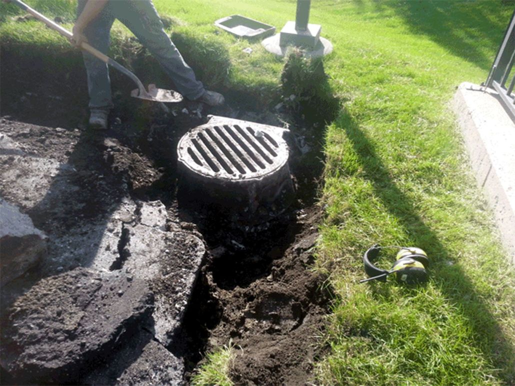 Worker digging around a round storm drain in a grassy yard with asphalt and soil exposed