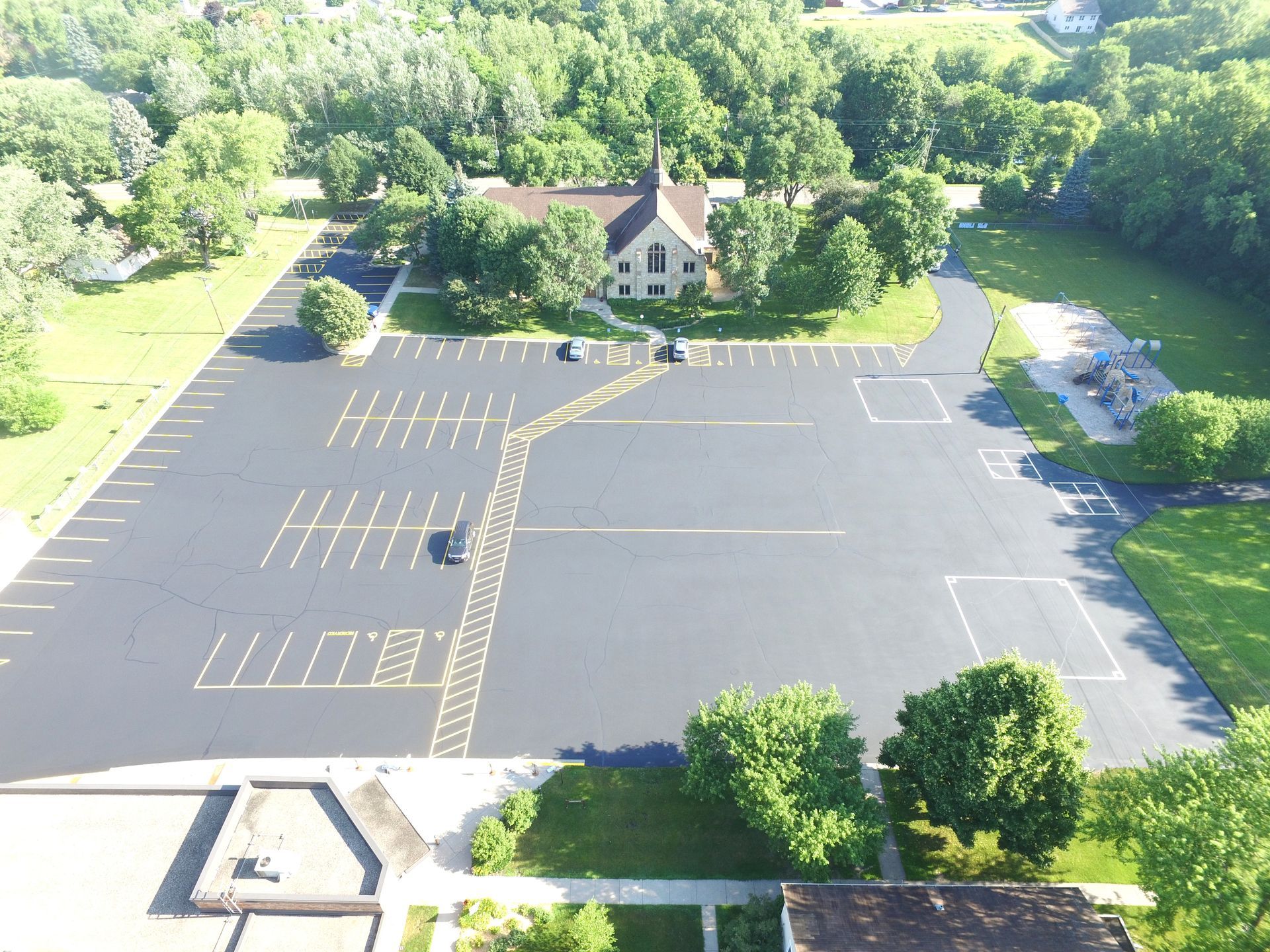Aerial view of an empty parking lot beside a small church, surrounded by trees and greenery.
