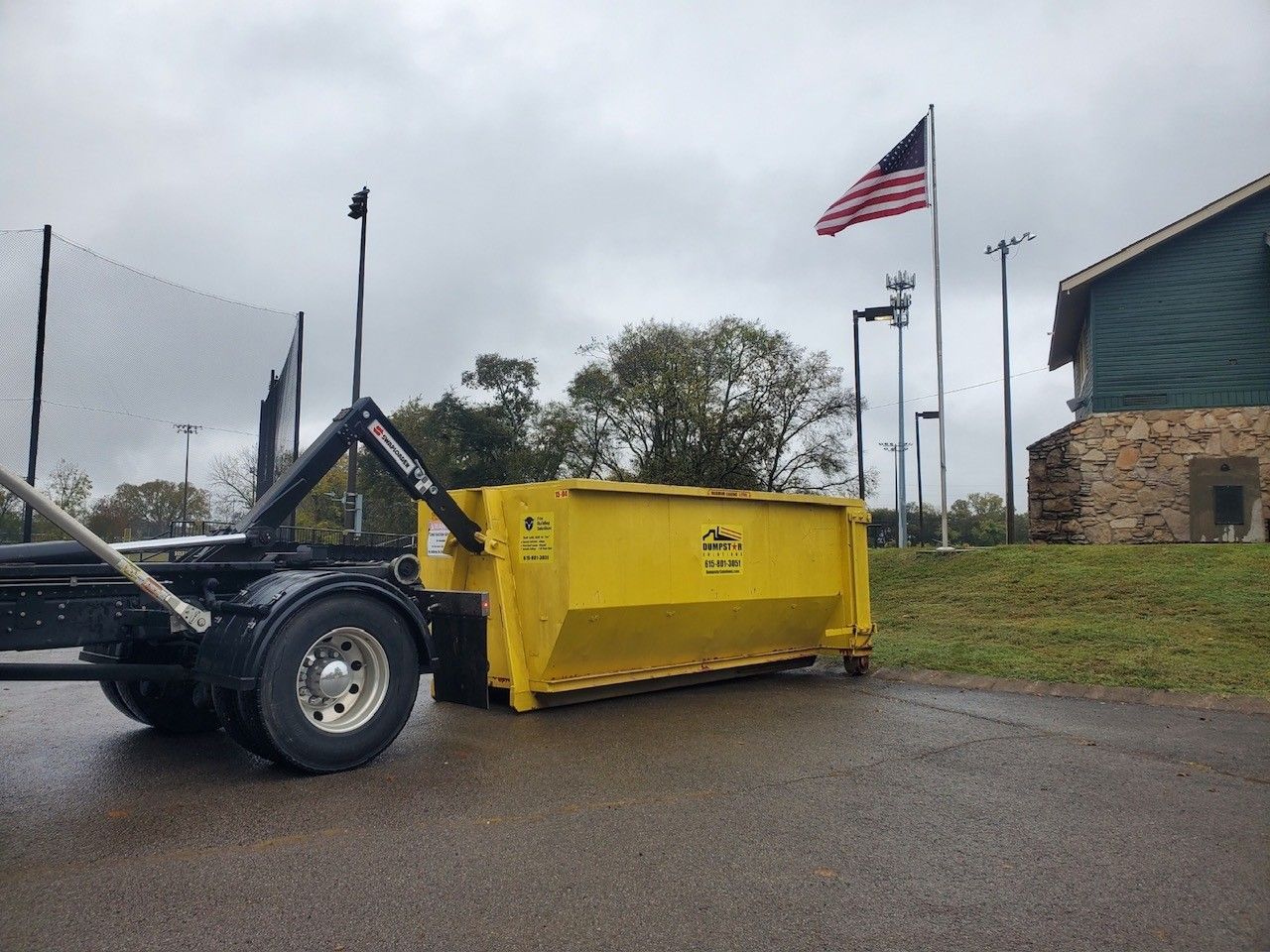 Yellow dumpster on a truck bed parked on asphalt near a building and American flag. Overcast sky.