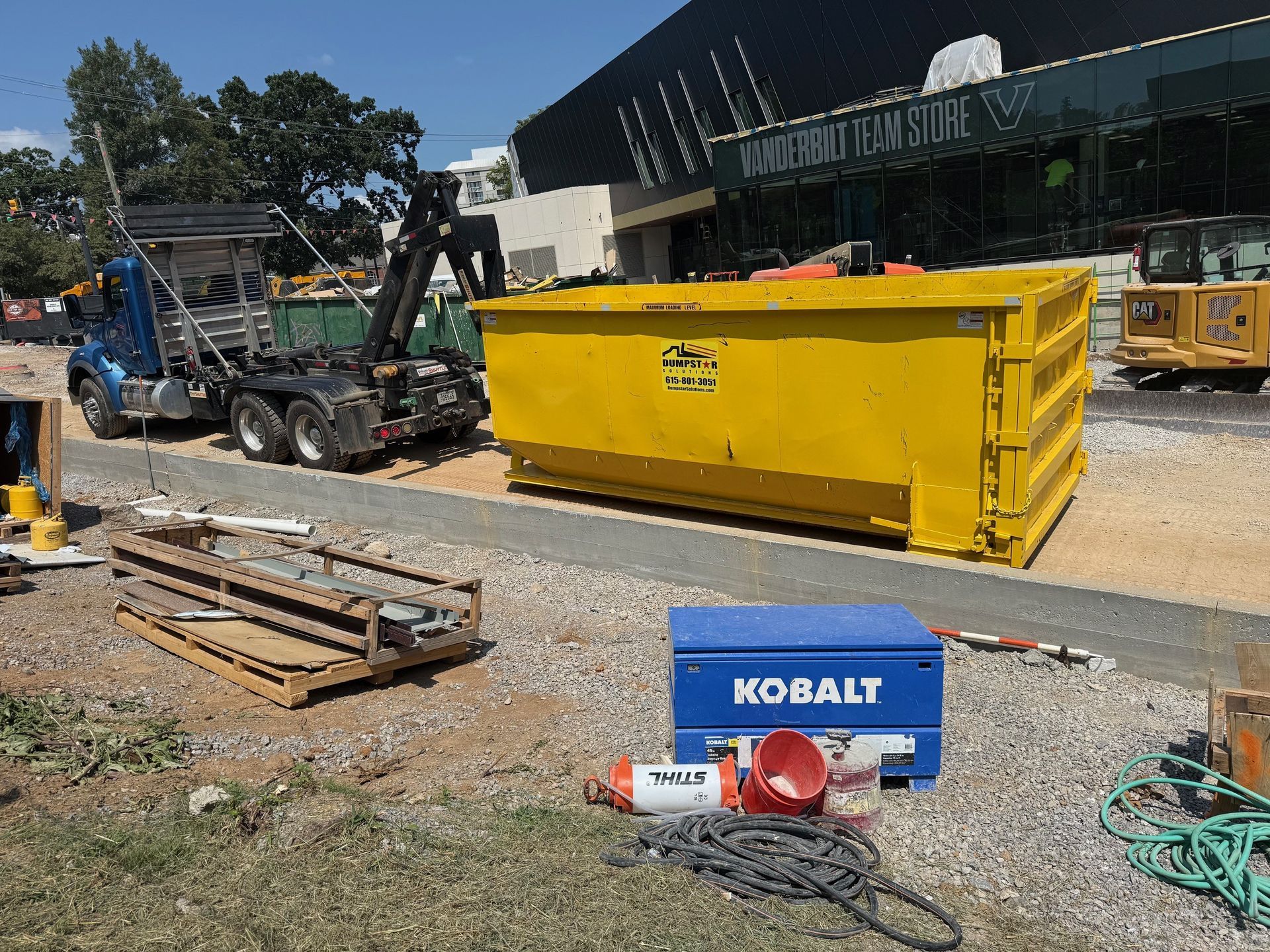 Yellow dumpster being loaded by truck at a construction site. Blue generator and tools in foreground.