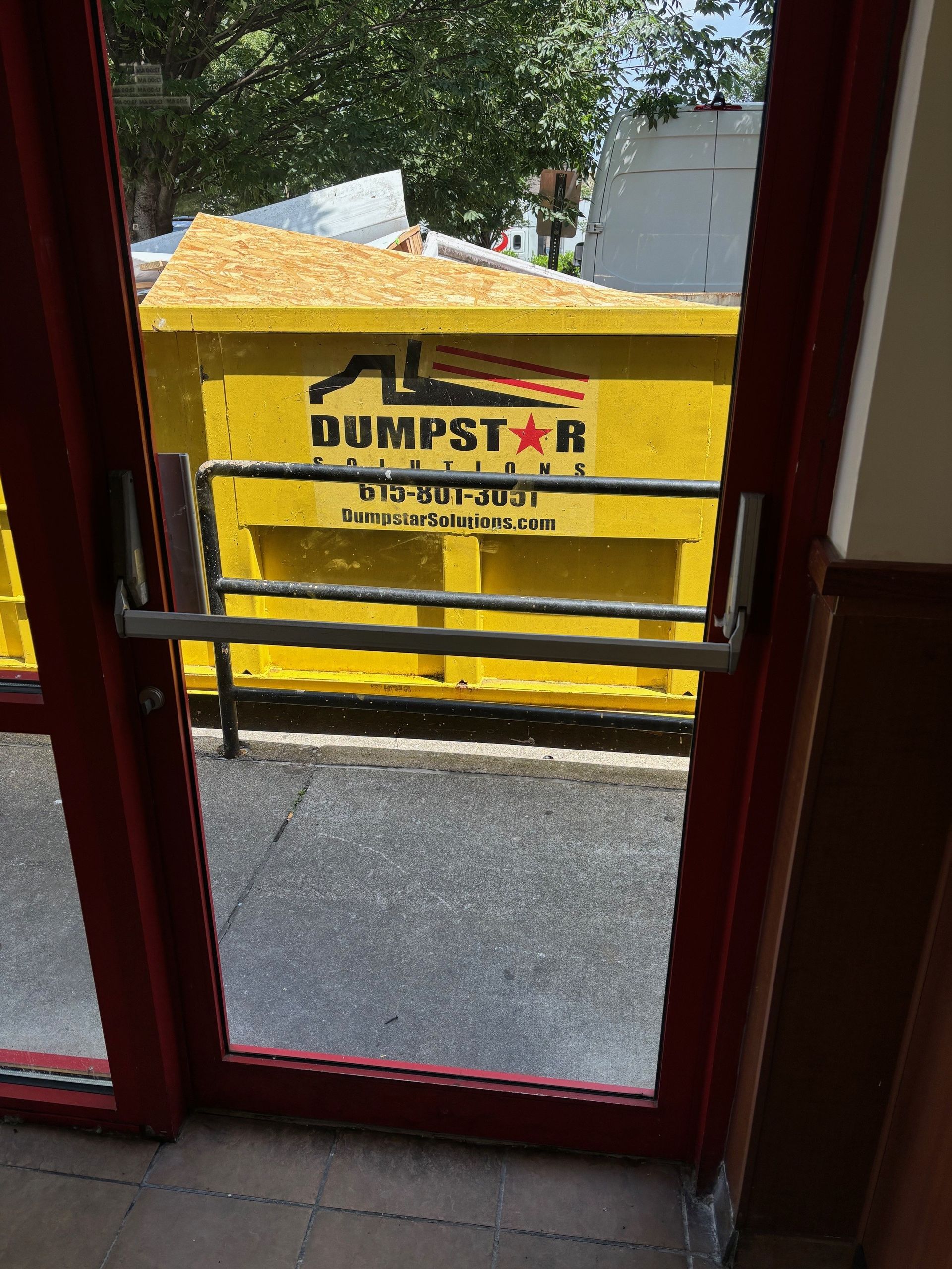 A yellow dumpster outside a door with a horizontal metal bar, red door frame.