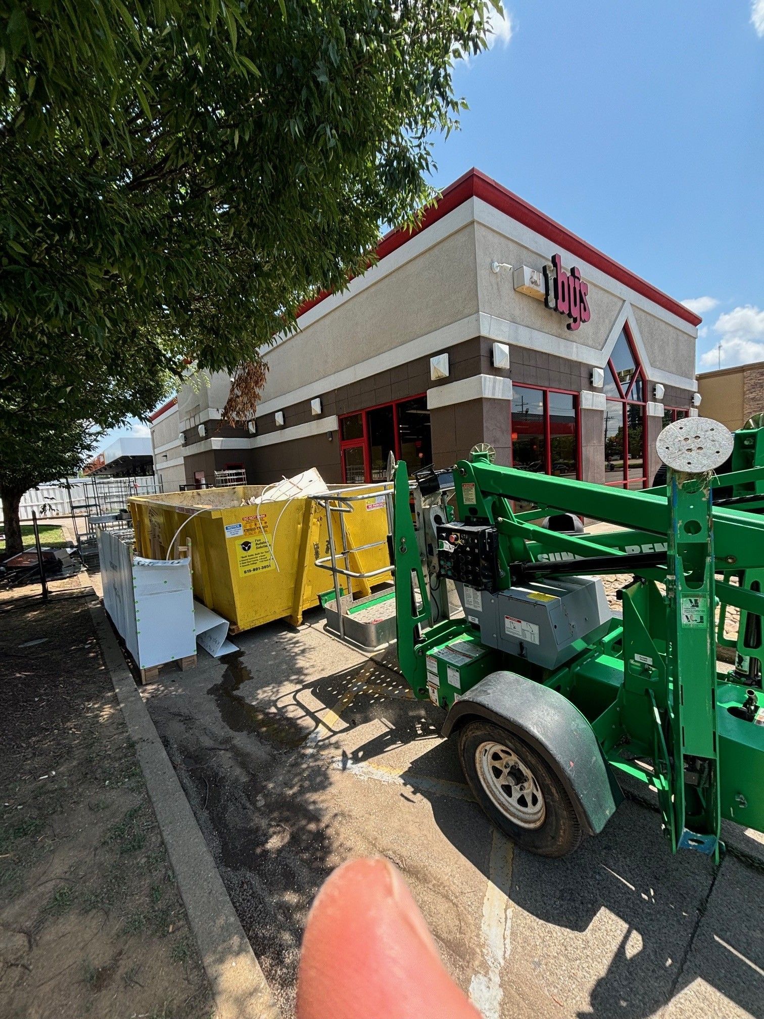 Exterior of a store with construction yellow dumpsters, green lift, and a red and white building facade.