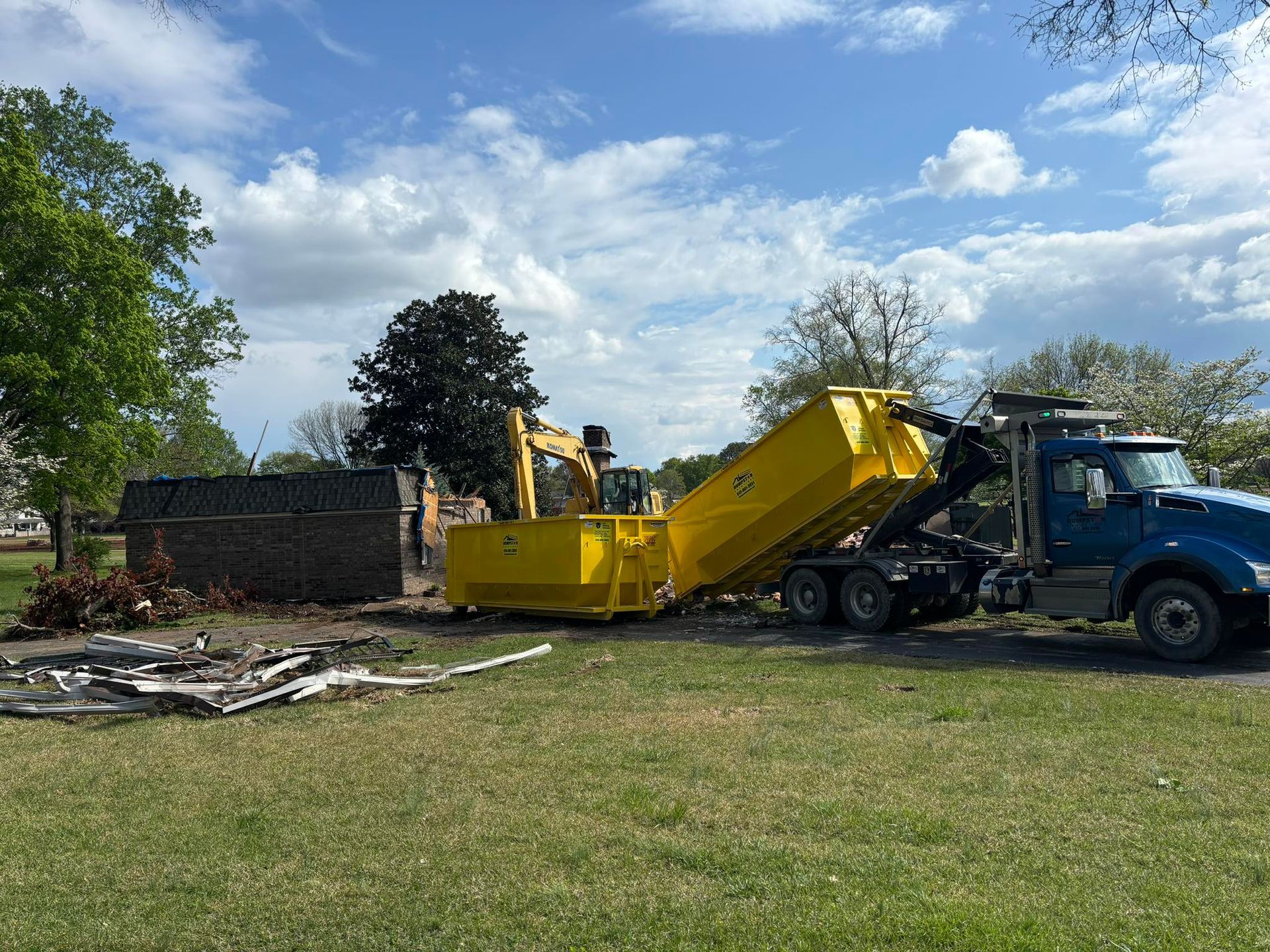 A demolition scene with an excavator and a yellow dumpster being loaded by a blue truck in a grassy area.