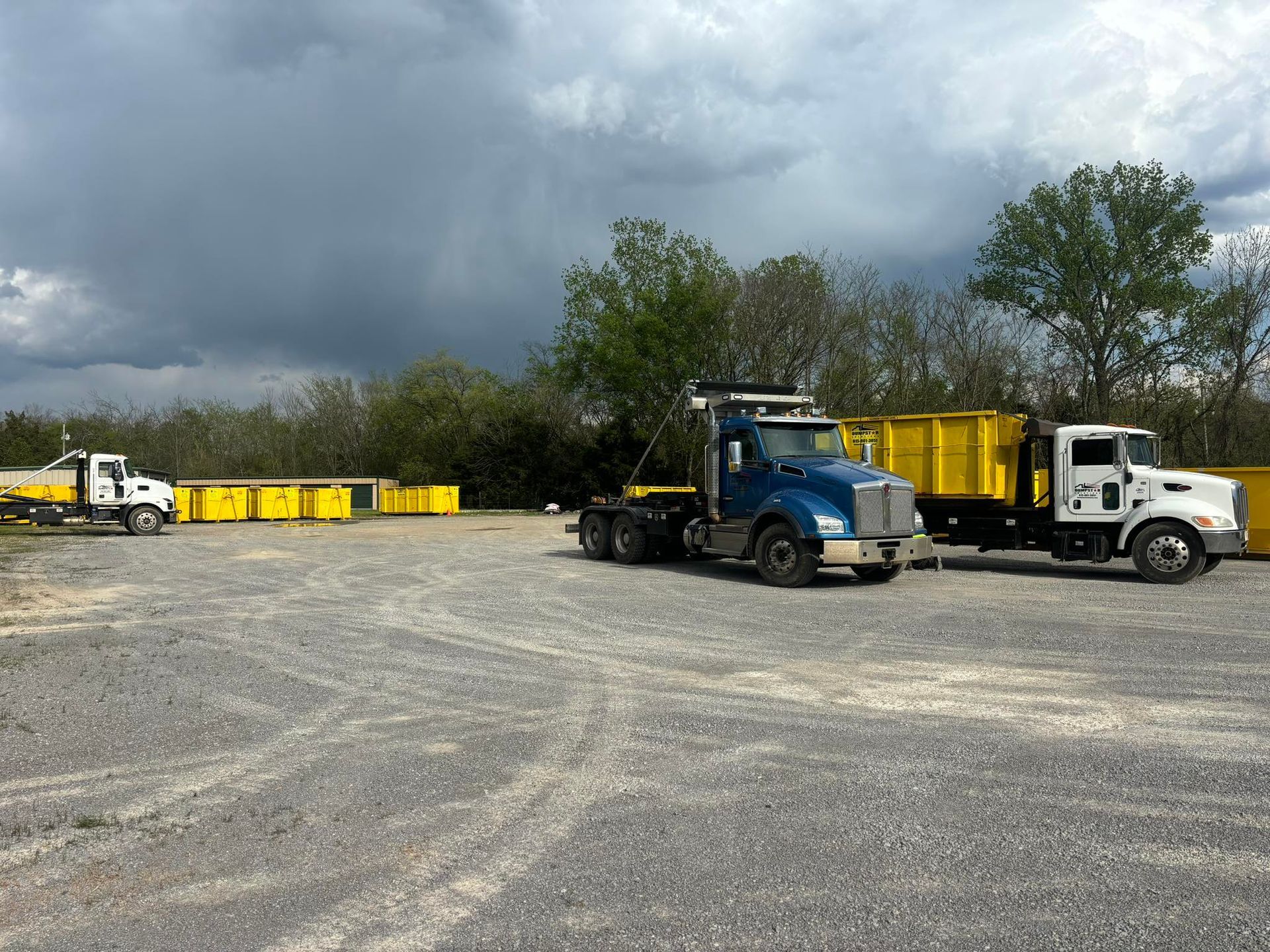 Three trucks, yellow dumpsters on gravel, dark sky.