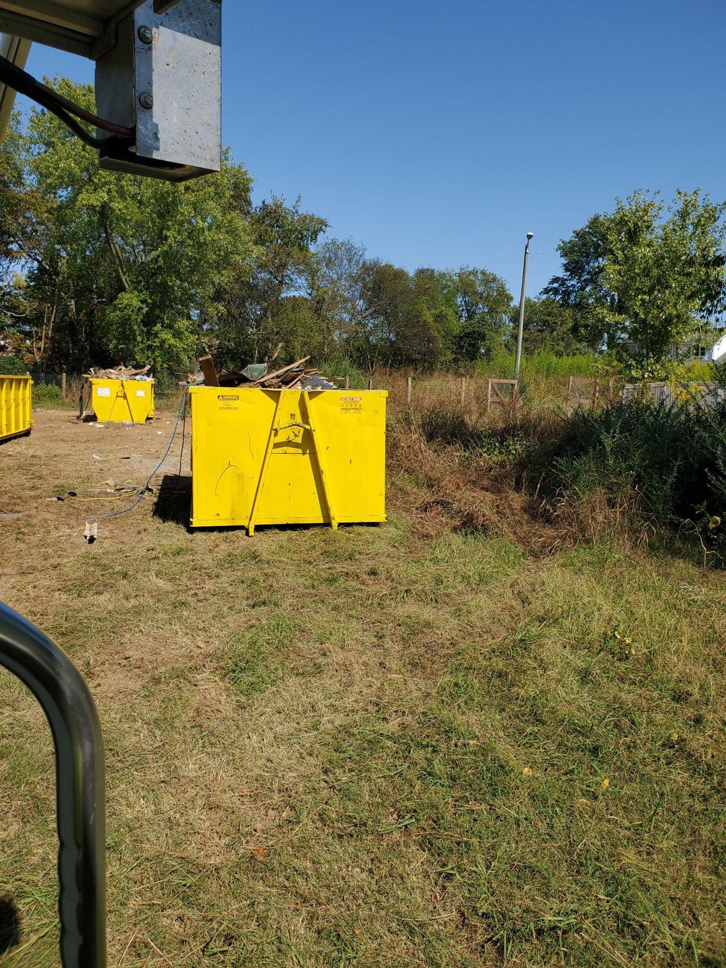 Yellow dumpsters on a grassy area, trees in background, clear blue sky.
