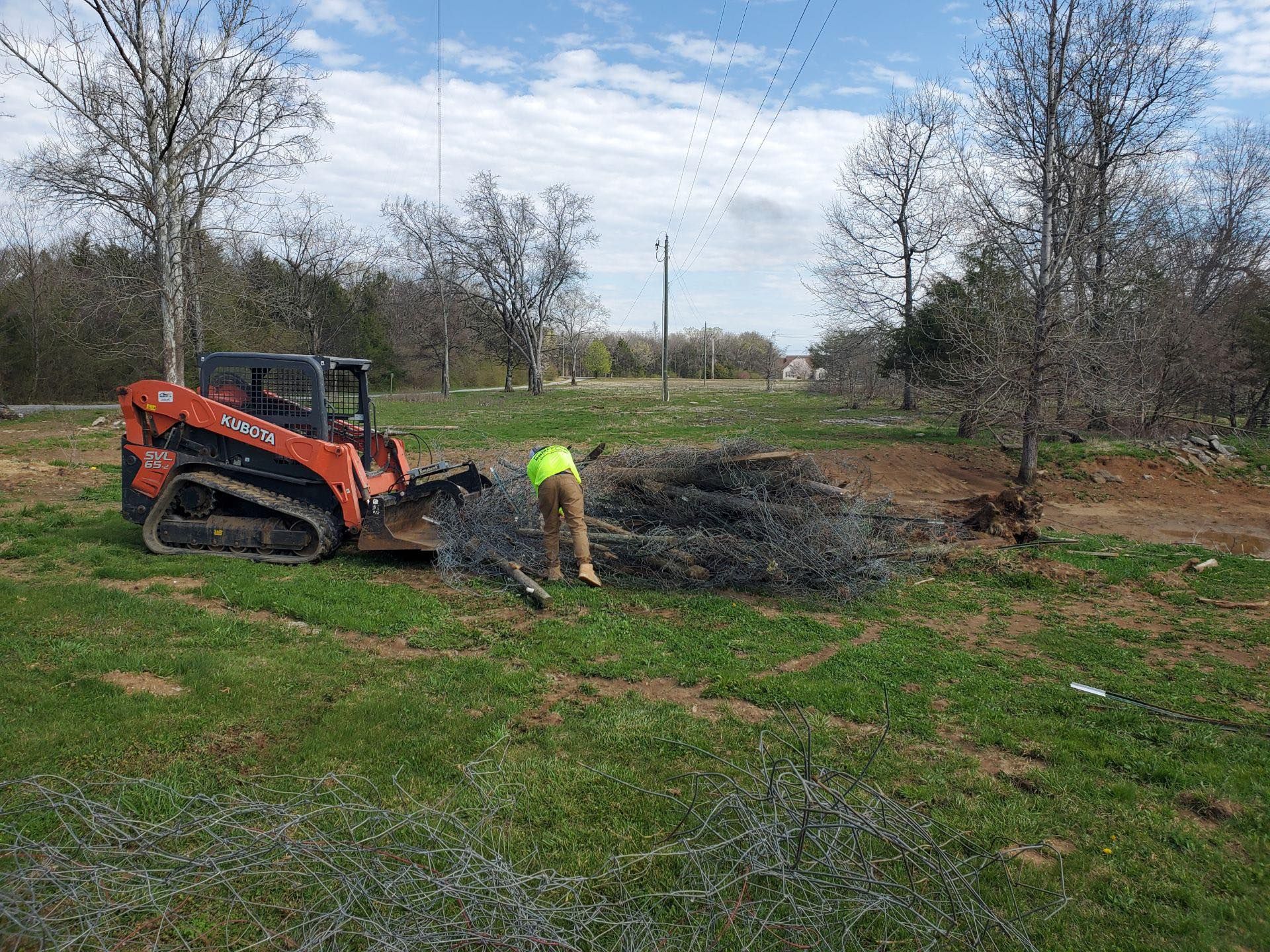 A worker loads barbed wire into a skid steer on a grassy field with trees and a blue sky.