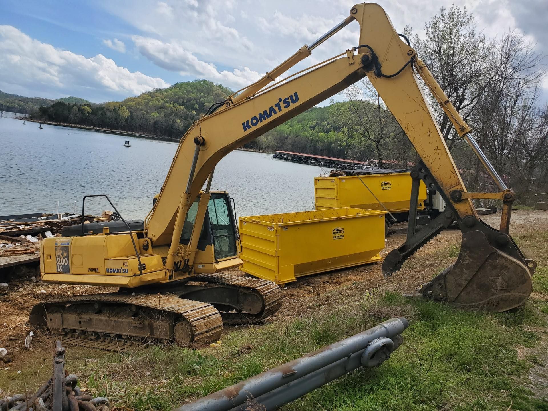 Yellow Komatsu excavator with bucket, next to yellow containers on the edge of a lake, surrounded by trees.