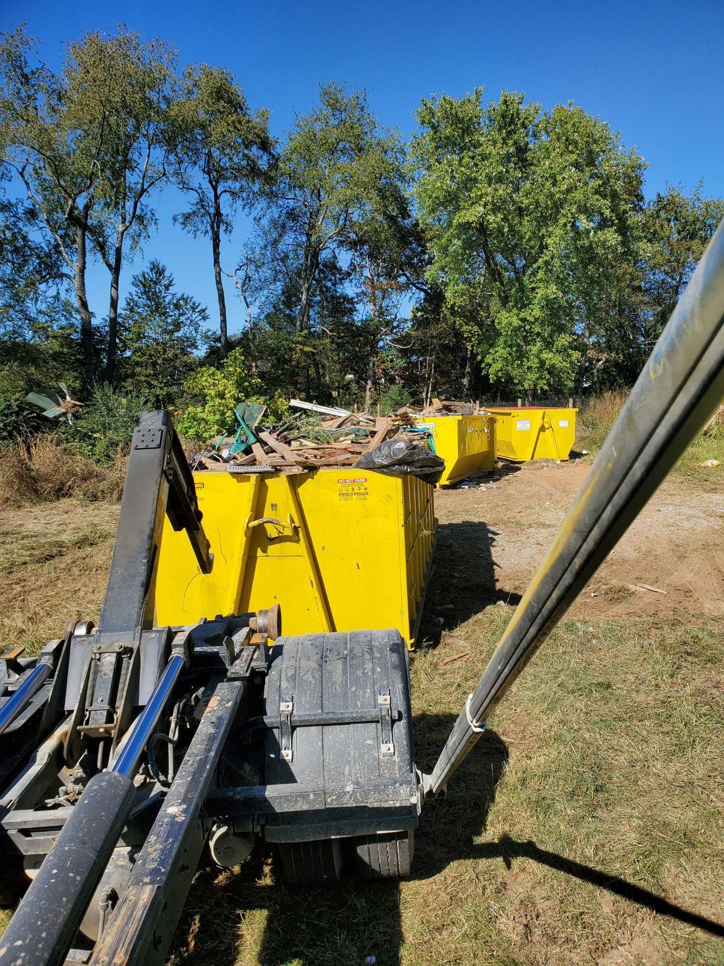 Yellow dumpsters full of debris in a sunny outdoor setting. Machinery in foreground, trees in the background.