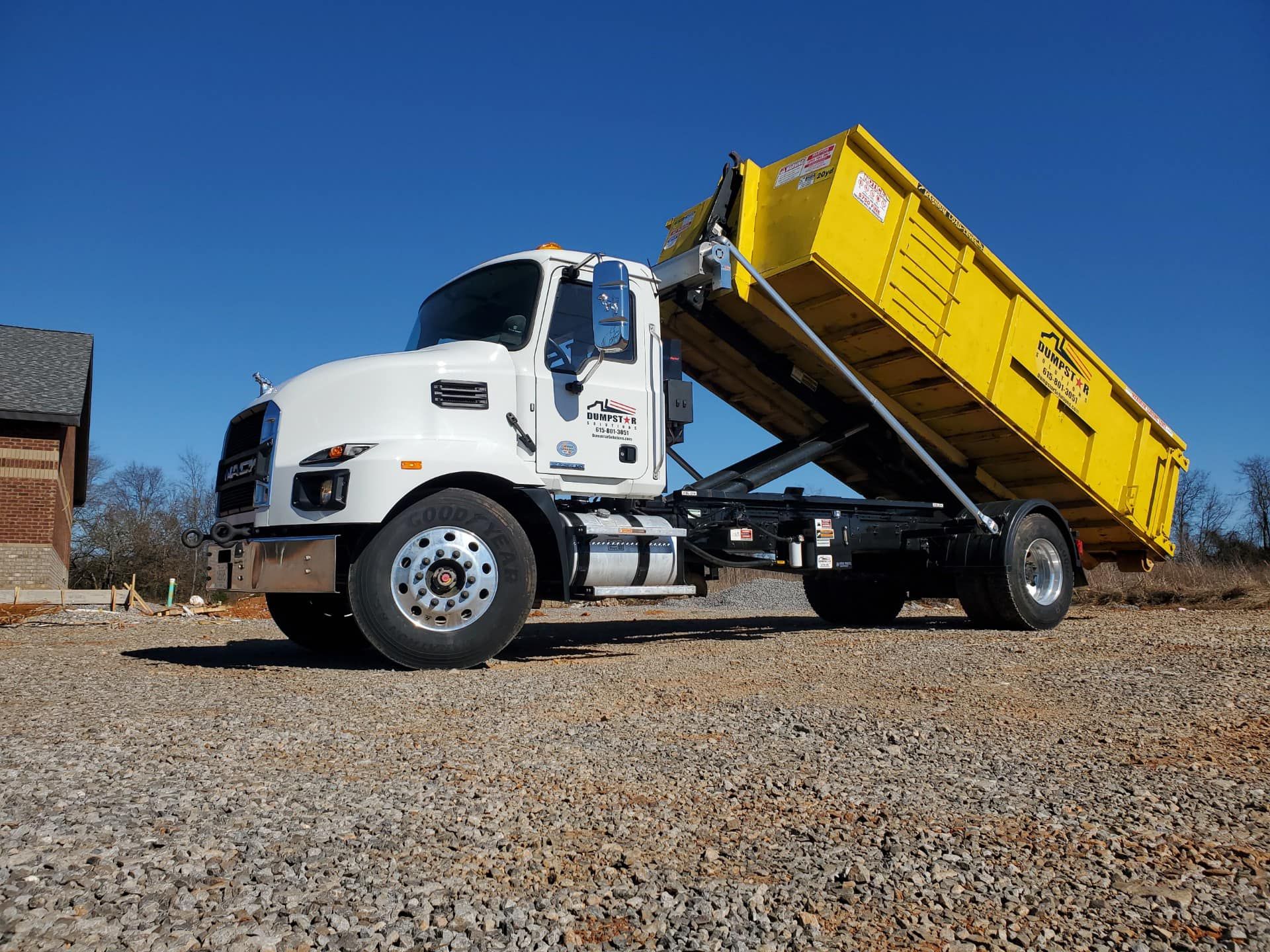 White dump truck with yellow container, tilted up, on gravel in daytime.