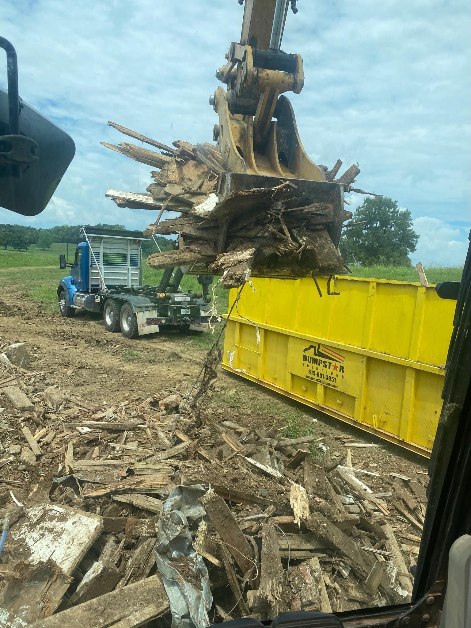 Excavator loading debris into a yellow dumpster next to a blue truck in a field.