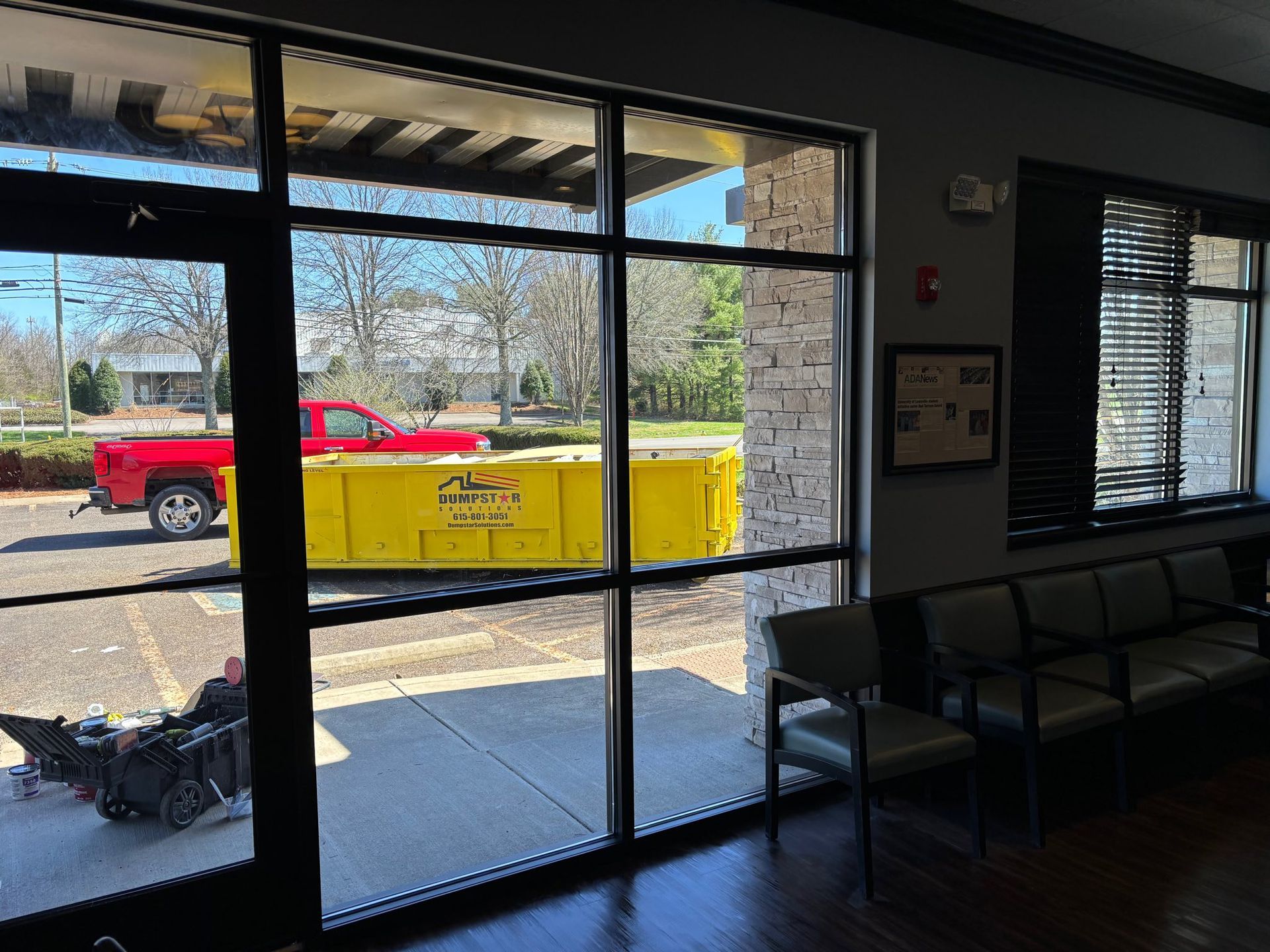 Interior view through glass windows; red truck and yellow dumpster outside. Chairs inside a waiting area.