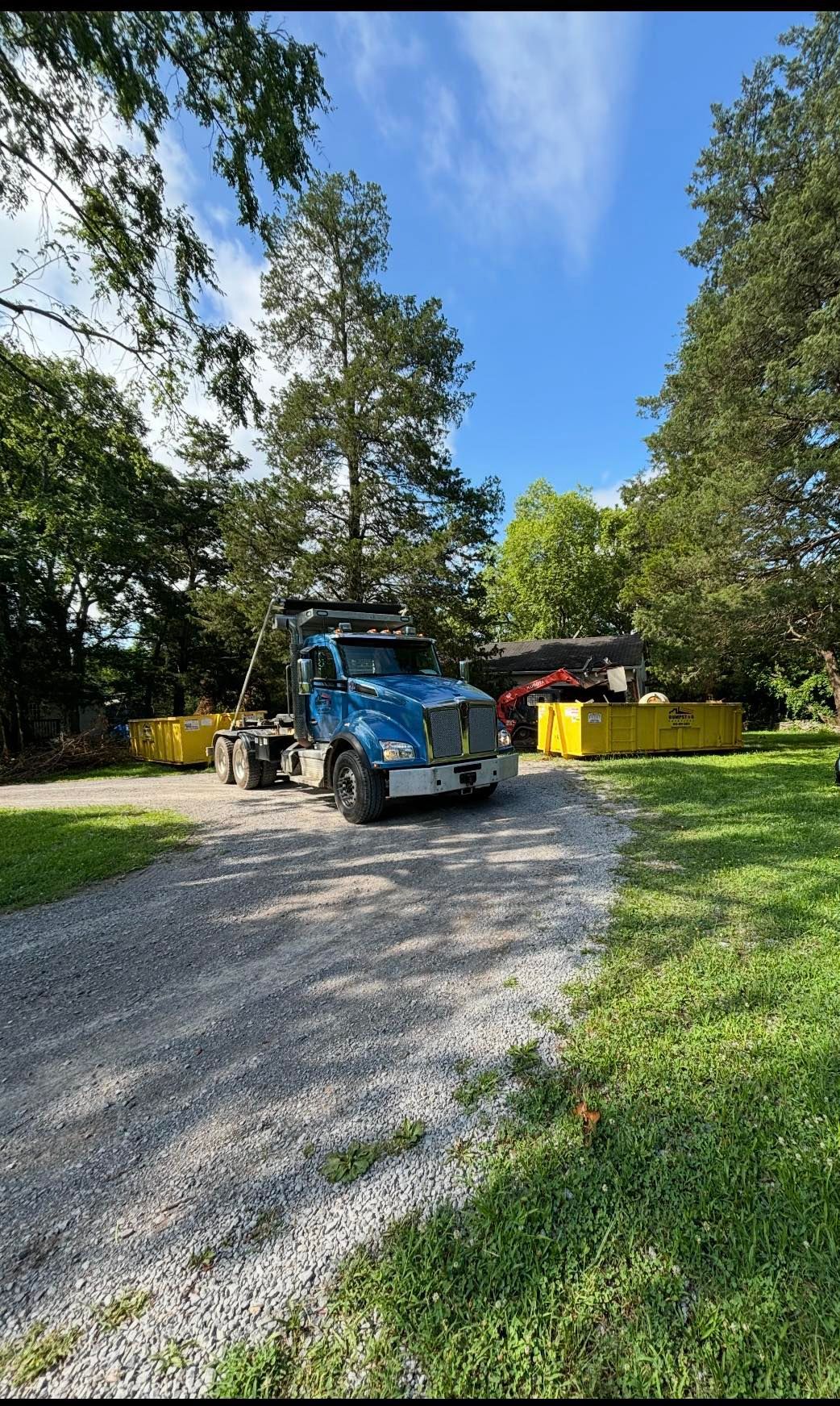 Blue truck on gravel driveway, beside yellow dumpsters in a wooded setting.