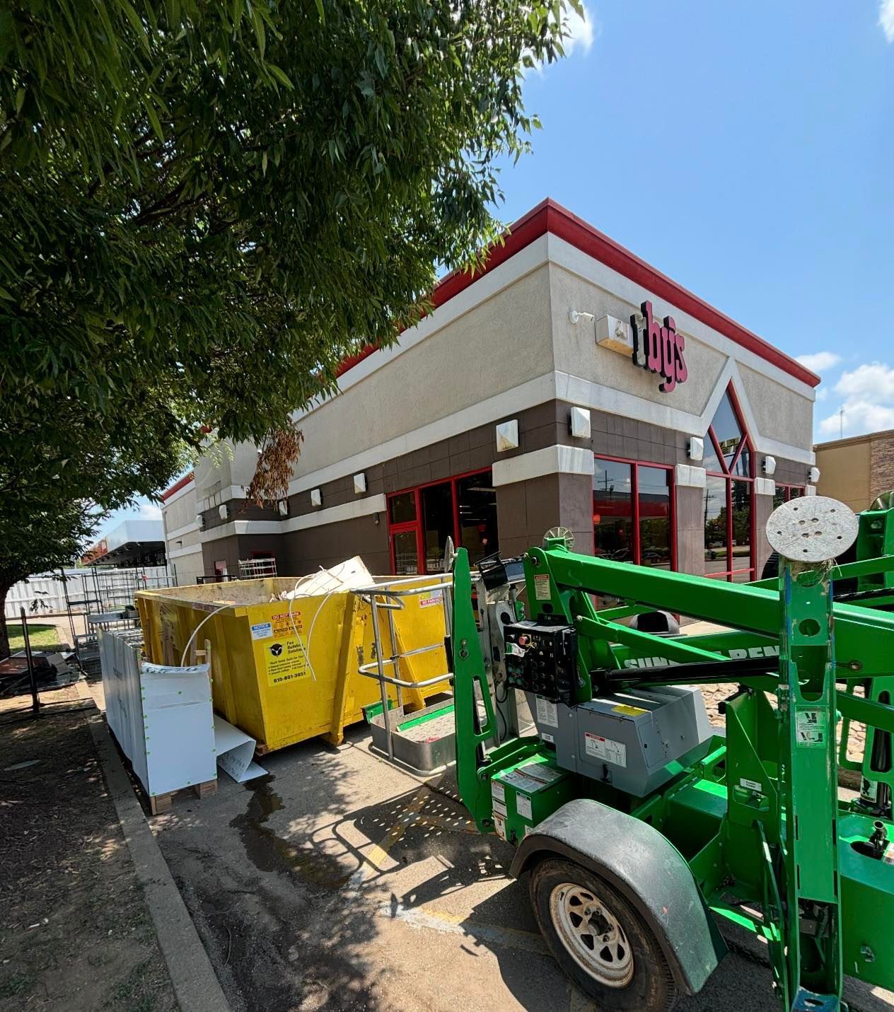 Exterior of a restaurant being renovated. Yellow dumpsters and green lift equipment sit outside under a bright blue sky.