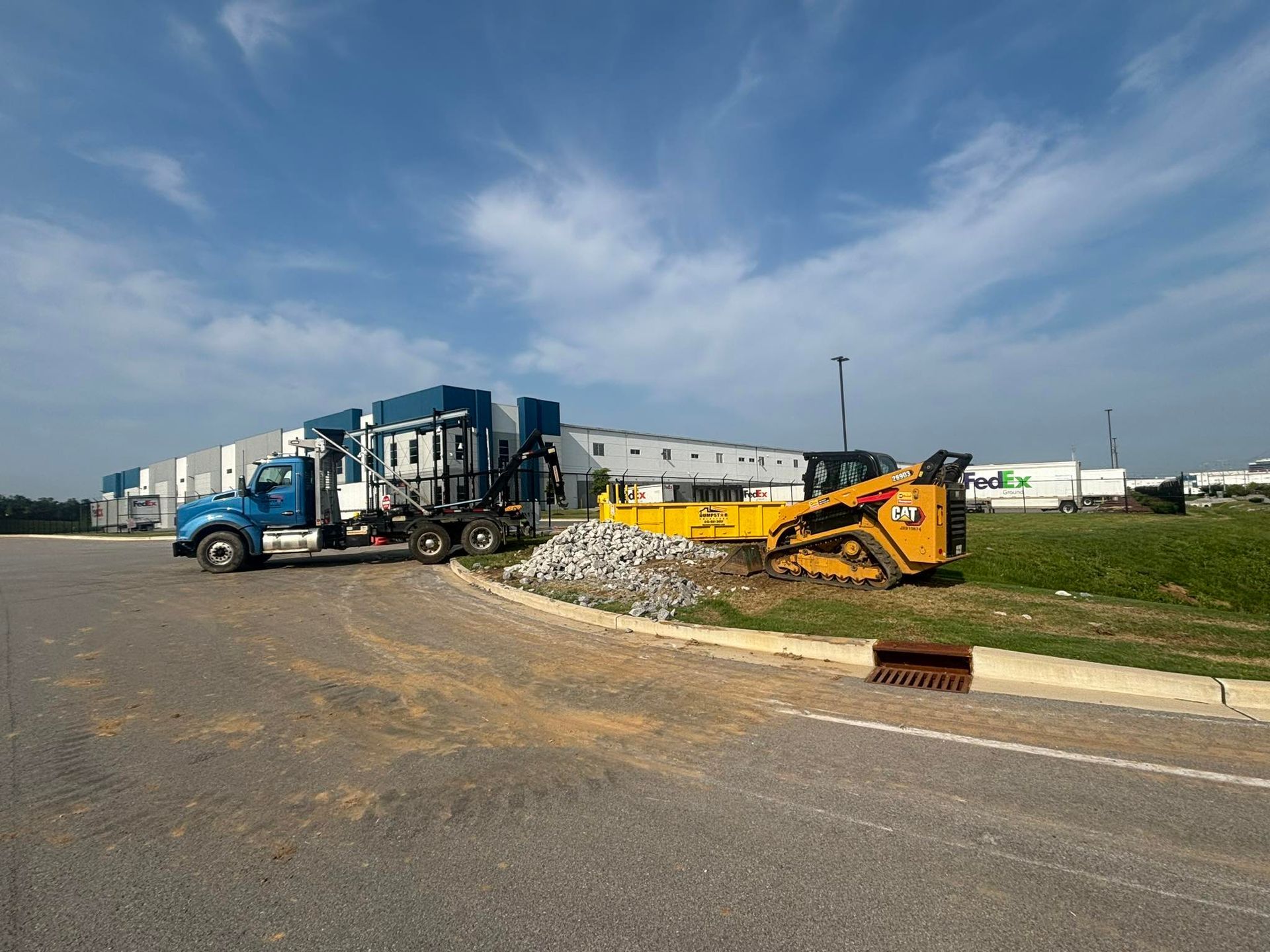 Blue truck and yellow skid steer on a construction site near a commercial building.